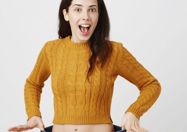 Astonished and happy young lady being impressed and excited because of losing weight, showing empty space in jeans by stretching it, standing over gray background. Girl is ready for summer.